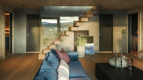 Moderne, svevende trapp i lyst treverk med glassvegg, sentralt plassert i en stue. Trappen gir et luftig uttrykk og leder opp til andre etasje over en blå sofa.