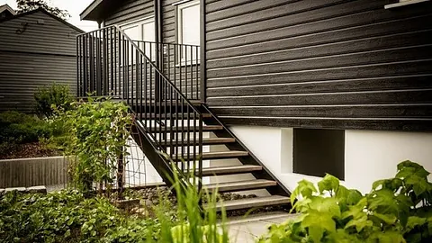 Utvendig ståltrapp med rekkverk montert på en bolig med mørk trekledning. Trappen leder ned til hagenivå og er omgitt av grønne planter.